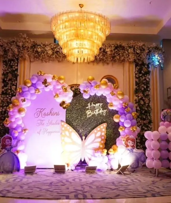 A stunning stage setup in a large banquet hall for a butterfly-themed birthday. The grand chandelier complements the purple and gold balloon arch and custom backdrop, creating a luxurious feel.