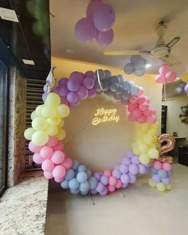 A beautiful pastel rainbow theme for a second birthday. This circular balloon arch uses soft shades of pink, yellow, purple, and blue, complemented by a 'Happy Birthday' neon sign.