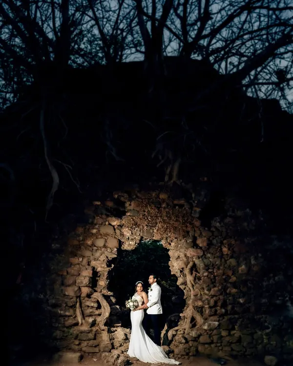 A wide, dramatic shot of Karen and Abhijeet within a stone ruin at twilight. The single light source creates a moody, cinematic feel, highlighting them against the dark woods.
