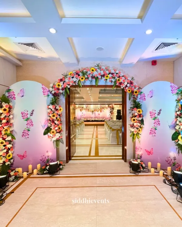 This enchanting entrance sets a dreamy tone for a 'Phoolon ki Haldi'. A grand floral arch with sunflowers and roses welcomes guests, while the pastel-hued backdrops with delicate butterfly motifs create a fairytale-like atmosphere, perfectly illuminated by soft floor lighting.
