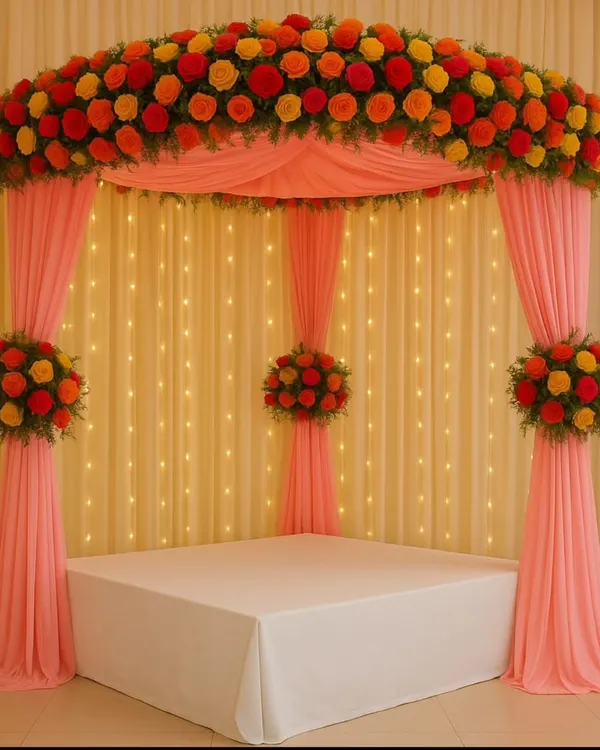 A classic mandap for Navratri or a wedding ceremony. This setup features a canopy of pink drapes topped with a vibrant arch of red, orange, and yellow marigolds, all illuminated by a backdrop of warm fairy lights.