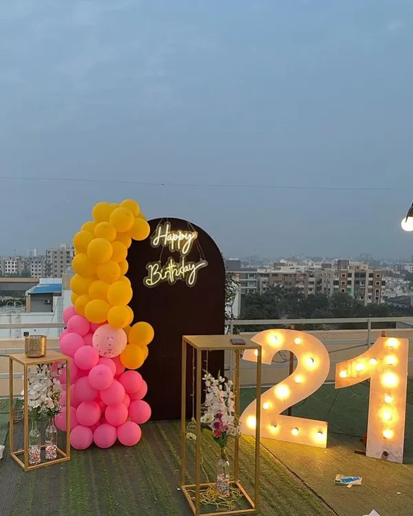 A chic 21st birthday decoration on a rooftop. This setup includes large illuminated marquee numbers, a "Happy Birthday" neon sign against a dark arch, and a bright pink and yellow balloon arrangement.