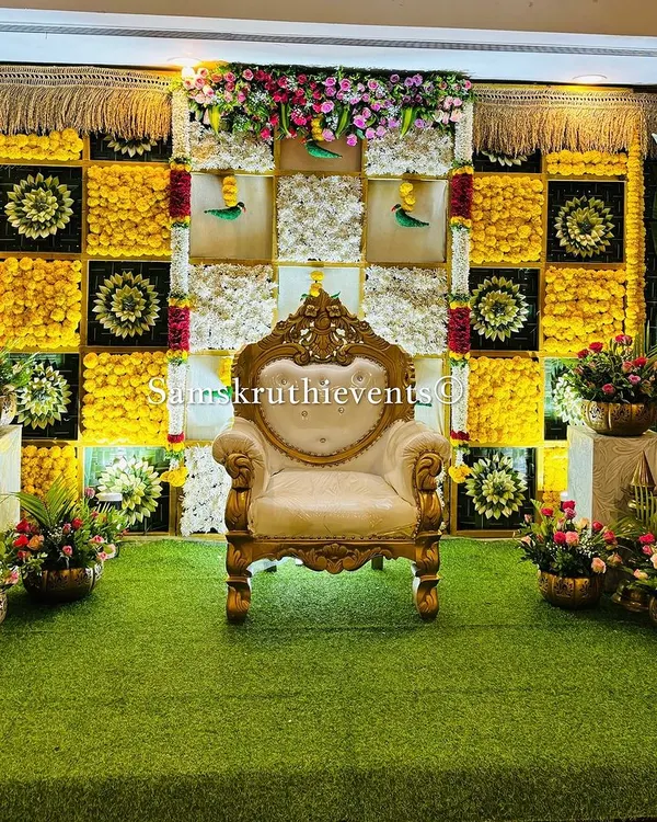 A classic and elegant backdrop for a housewarming ceremony. The design features a checkerboard pattern of yellow marigolds and white chrysanthemums, accented with woven leaf details and large banana plants.
