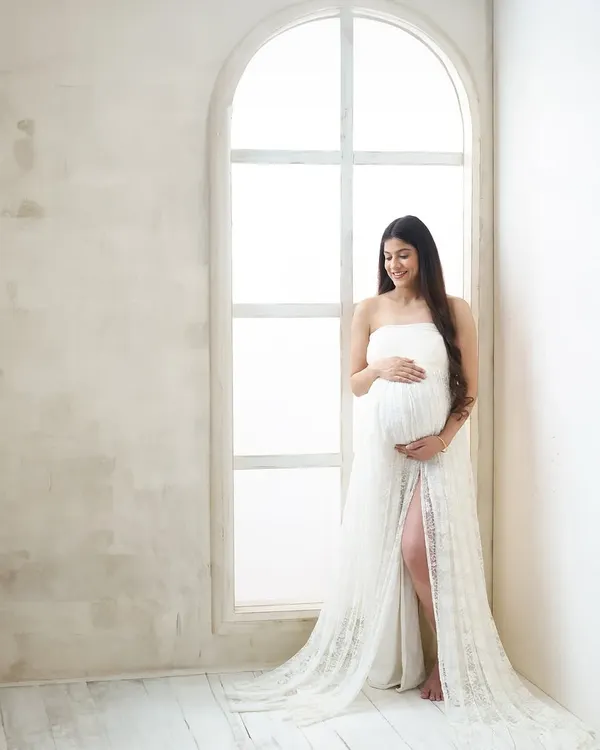 A full-length portrait using the beautiful natural light from an arched window. The bright, clean setting and elegant white lace gown create a serene and angelic atmosphere.