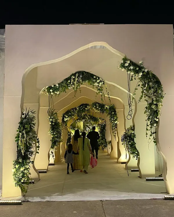 A modern and artistic entrance structure for an evening reception. The custom-built, free-flowing arched tunnel is accented with delicate white florals and uplighting, creating a sculptural and impressive welcome.
