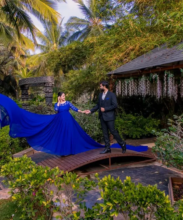 A couple on a wooden bridge surrounded by lush greenery, with the flowing blue dress adding a dramatic splash of color to the natural scene.