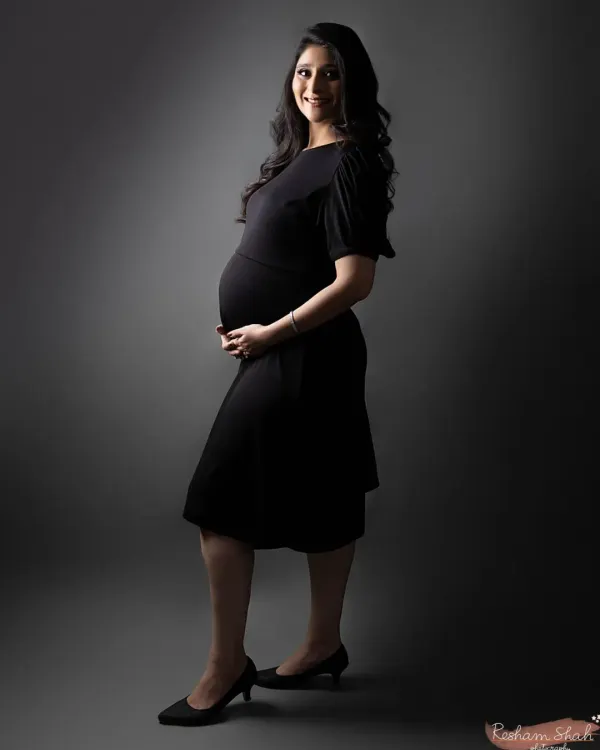 A full-length silhouette shot that beautifully defines the baby bump. The mom-to-be stands in a simple black dress against a grey backdrop, creating a timeless and elegant maternity portrait.