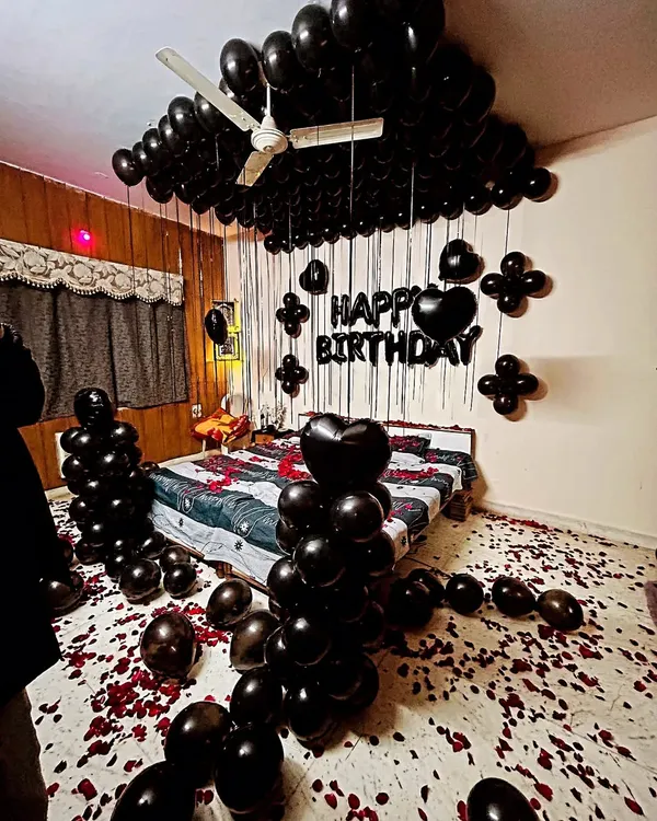 A dramatic and romantic all-black themed room decoration. This setup includes black balloons covering the ceiling, heart-shaped foil balloons, and rose petals on the floor and bed.