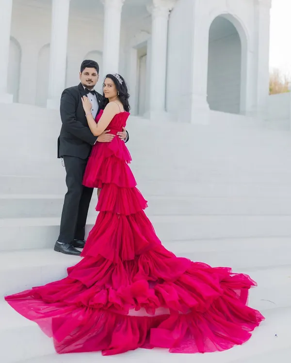 A cinematic portrait of an expecting couple on grand, white steps. The vibrant red of her tiered gown contrasts beautifully with the classic black tuxedo he wears, creating a powerful and romantic image that celebrates their journey to parenthood.