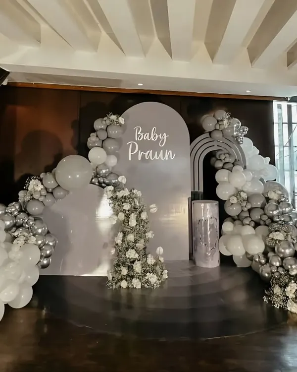 An elegant and sophisticated baby announcement or shower for "Baby Praun." The stage features a chic grey, white, and silver color palette with beautiful balloon arches and floral accents.