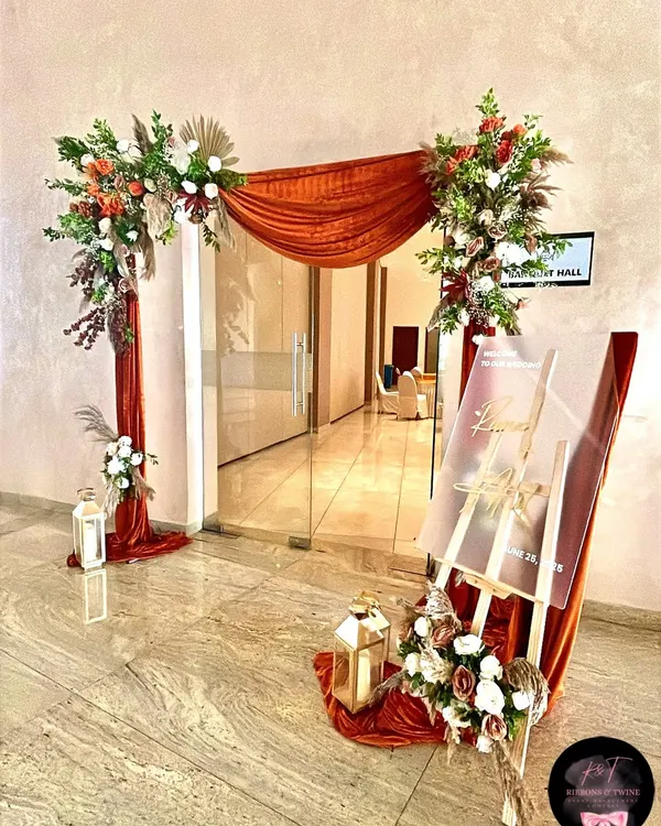 A grand entrance for a wedding hall, styled with an asymmetrical arch of rich fabric and florals, lanterns, and a custom acrylic welcome sign.