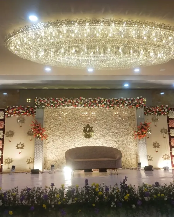 This is a wide view of a reception stage I designed, featuring a massive crystal chandelier overhead. The backdrop is a full wall of white flowers with a central Ganesha motif and lotus accents.