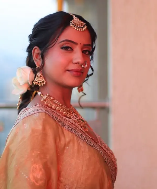 A beautiful portrait of a bride in a golden lehenga. Her hair is styled in a soft, romantic braid with a fresh white rose, perfect for a daytime wedding.