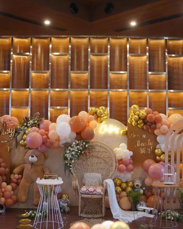 A full view of a 'We Can Bearly Wait' baby shower. The setup features a wicker peacock chair for the mom-to-be, surrounded by a cascade of balloons in earthy tones and a friendly teddy bear.