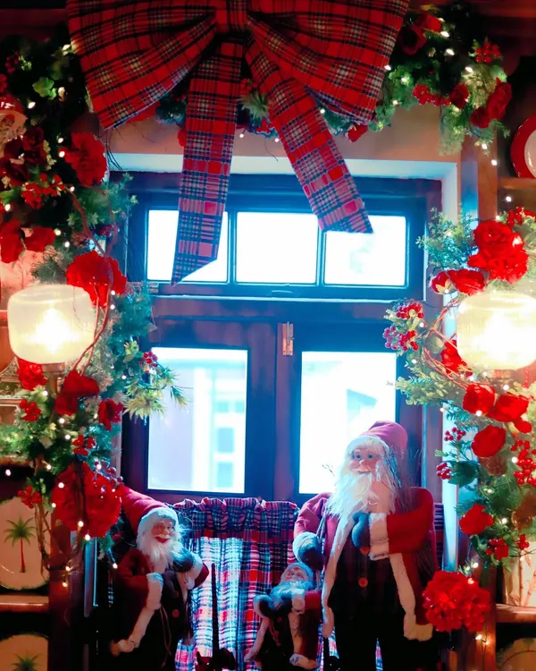 A classic Christmas window display for Scarlett House, featuring Santa figures, a large plaid bow, and a lush garland adorned with red flowers and fairy lights.