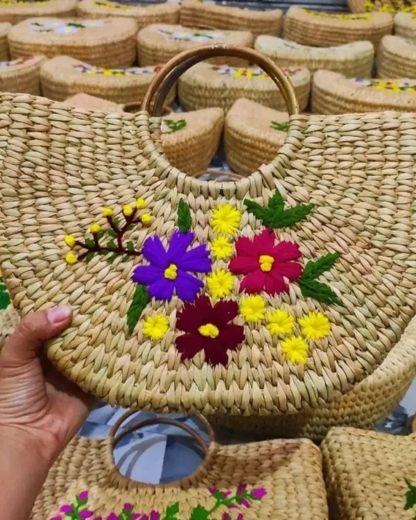 A closer look at one of our fan-shaped sabai grass bags, featuring colorful and detailed floral embroidery. Held in hand, you can see the fine craftsmanship and sturdy wooden handle that make this artisan bag both beautiful and practical.