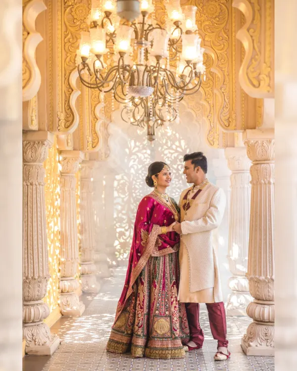 A regal moment captured in a palace-like corridor. The couple stands under a grand chandelier, with light filtering through ornate pillars, creating a timeless and cinematic portrait of their love story.