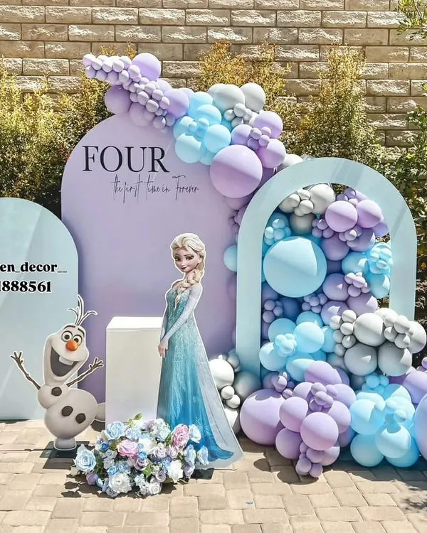 An elegant outdoor Frozen-themed party setup for a fourth birthday. The design features Queen Elsa and Olaf cutouts, a beautiful balloon cascade in pastel purple and blue, and custom arched backdrops for a sophisticated look.