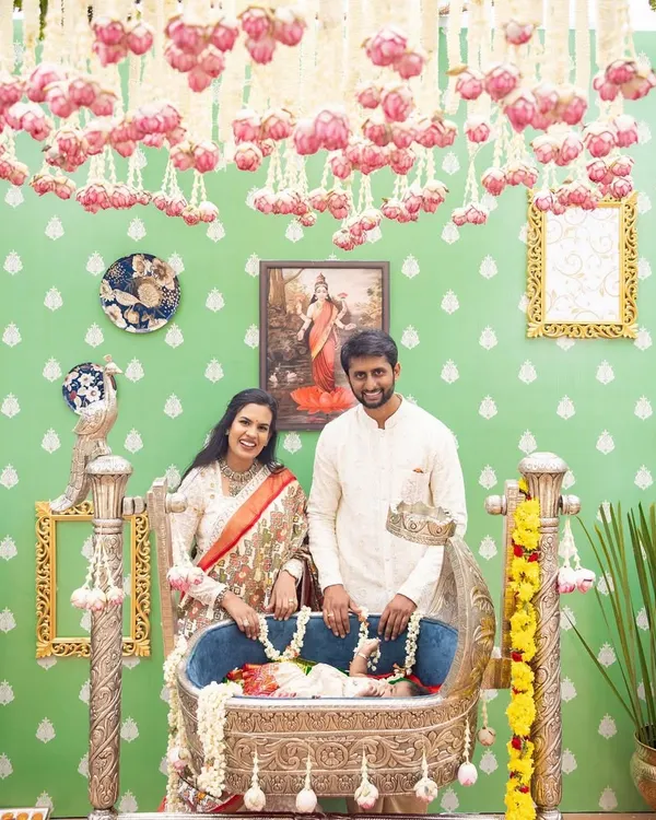The happy family at their daughter Tridha's naming ceremony. The decor features a beautiful silver cradle and a backdrop inspired by Ravi Varma paintings.