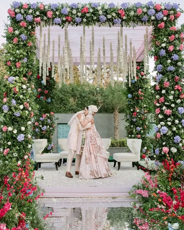 A beautiful moment of embrace under a lush floral mandap. The structure is covered in pink, purple, and red flowers, with hanging tuberoses adding a traditional touch to the modern pastel theme.