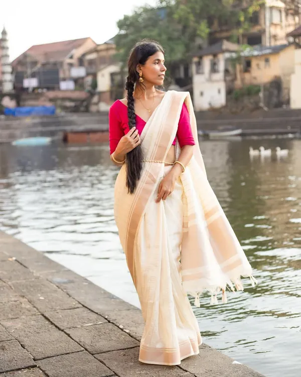 A poised, full-length shot of a classic off-white and pink silk saree, showcasing its elegant drape and texture against a scenic riverside backdrop.