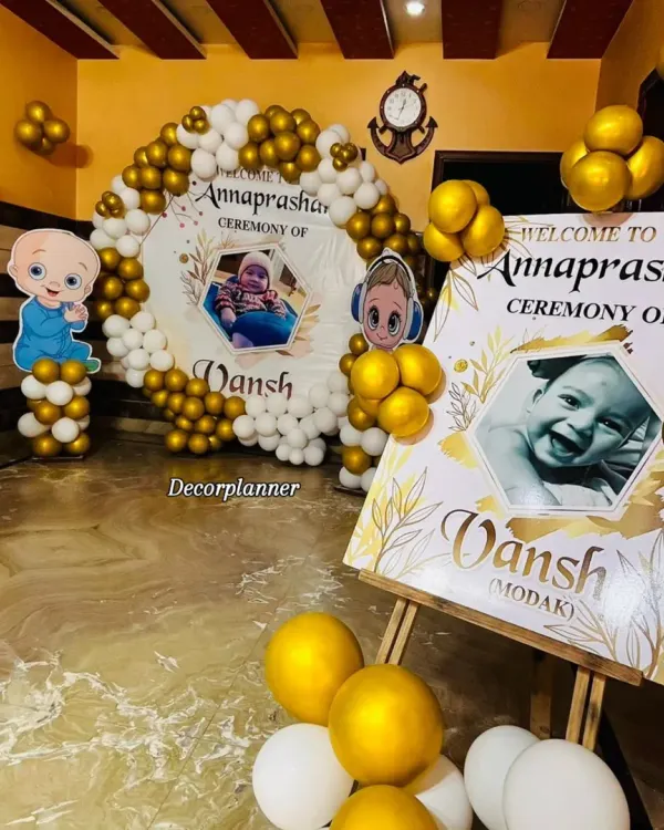 A complete view of the personalized Annaprashan ceremony for baby Vansh. The setup includes a custom welcome easel and a matching circular backdrop, both featuring his photo, framed by a beautiful white and gold balloon arch.