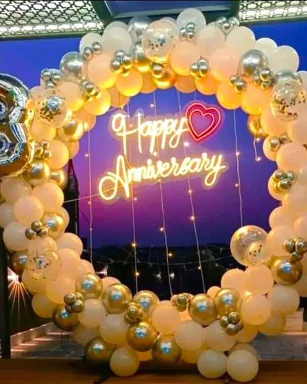 A beautiful anniversary decoration featuring a balloon ring in white, gold, and transparent confetti balloons. The 'Happy Anniversary' neon sign and fairy lights create a warm and celebratory glow.