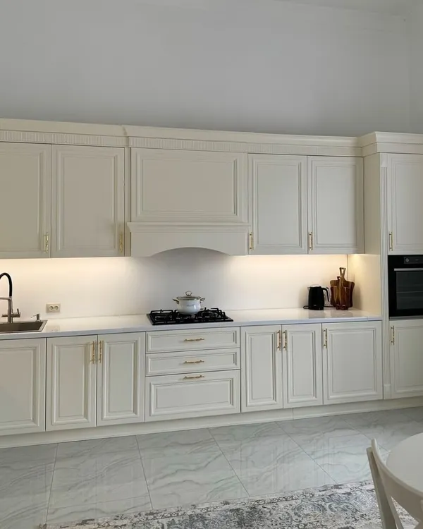 A beautiful, expansive kitchen in a classic off-white finish. The moulded cabinet doors, elegant gold handles, and integrated range hood cover create a seamless and timeless look of luxury.