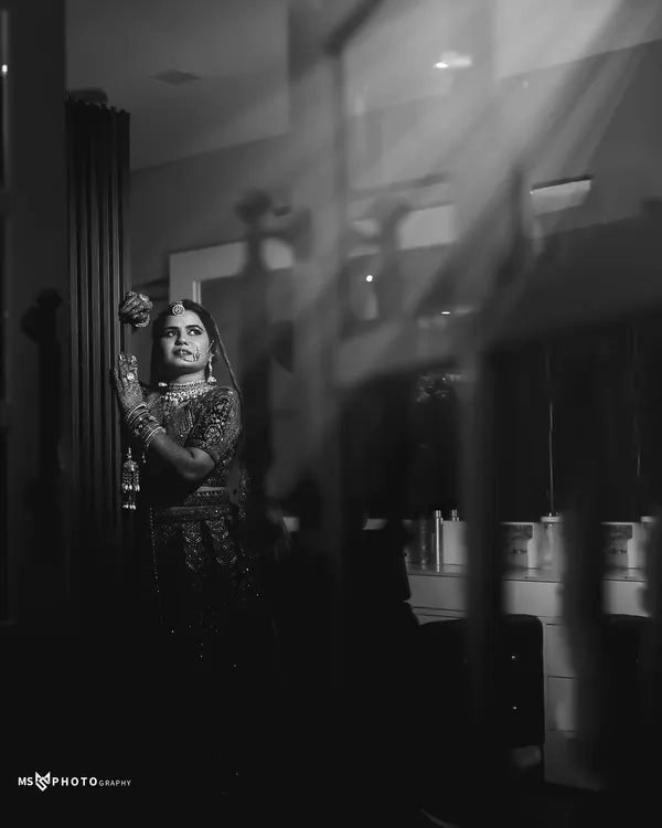 A moody and artistic black and white bridal portrait. The bride is captured through a reflection, with dramatic light and shadows creating a sense of quiet contemplation and timeless elegance.