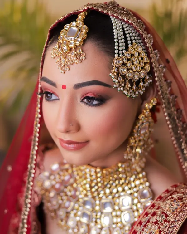 A quintessential bridal portrait focusing on flawless HD makeup. The model's red eyeshadow and defined features are perfectly complemented by heavy kundan jewellery against a traditional red bridal dupatta, creating a classic and aspirational look for any MUA's portfolio.
