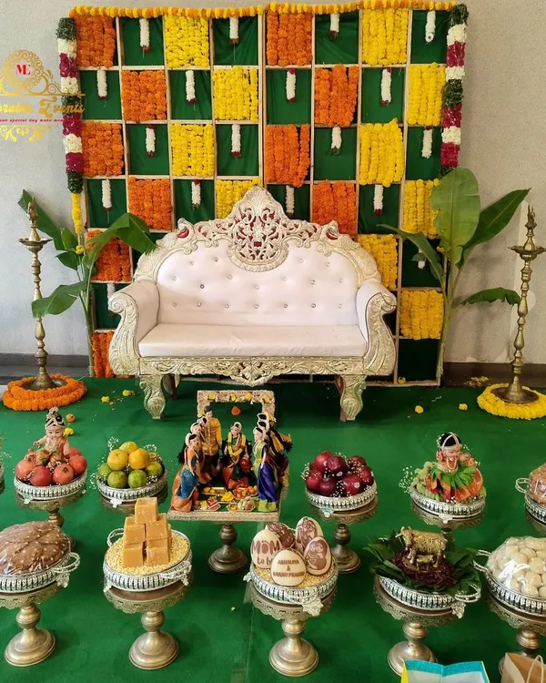 A traditional setup for a baby shower or naming ceremony, featuring a backdrop of marigold flowers and banana leaves, an ornate sofa, and brass lamps (diyas).