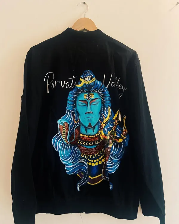 This is the finished Lord Shiva bomber jacket, hung to display the full artwork clearly. The vibrant blues and golds stand out against the black fabric, creating a visually stunning piece.