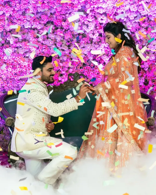 A picture perfect engagement moment. The groom is on one knee, surrounded by a cloud of smoke and a shower of confetti, against a stunning floral backdrop.