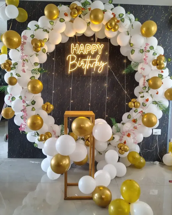 An elegant and classic birthday backdrop featuring a full circle balloon garland in white and gold. The setup is enhanced with a 'Happy Birthday' neon sign and a small pedestal for the cake.