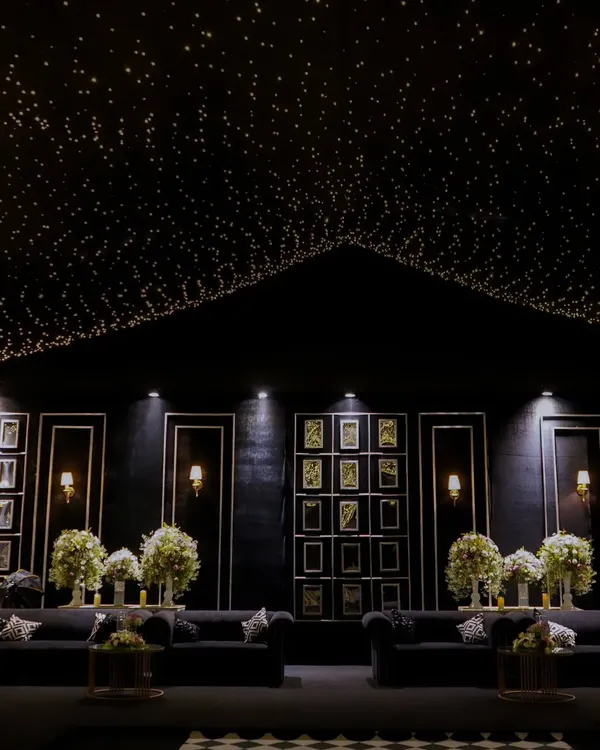 A wide view of a modern monochrome lounge we designed. A ceiling of countless fairy lights creates a starry night effect above a chic black seating area with a gallery wall, perfect for a sophisticated corporate mixer.