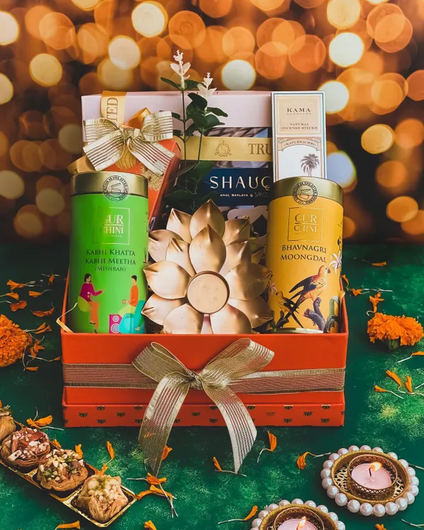 The "Raunaq" hamper is designed to bring splendor to any Diwali celebration. This vibrant orange box includes gourmet Indian snacks, natural incense sticks, and a stunning gold-plated lotus diya as the centerpiece.