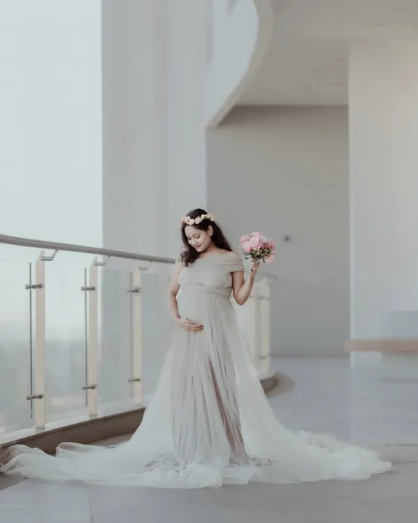 A full-length portrait of an expecting mother in a stunning, light-colored maternity gown, holding a bouquet of pink roses. This elegant maternity photo was taken in a bright, modern indoor location.