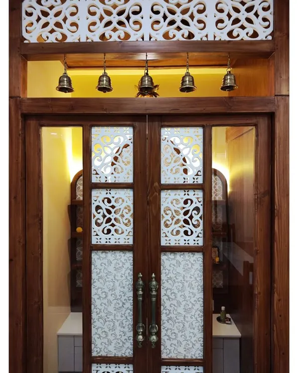 The doors to a divine space. This pooja room entrance features custom wooden doors with frosted glass, intricate jaali cutouts, and traditional brass handles. The transom window above with hanging bells adds a final touch of temple-inspired elegance.