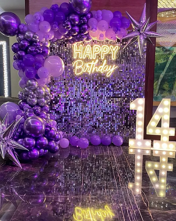 A glamorous 14th birthday setup with a purple shimmer wall. This decoration features a stunning organic balloon garland in shades of purple and silver, a 'Happy Birthday' neon sign, and large marquee numbers.