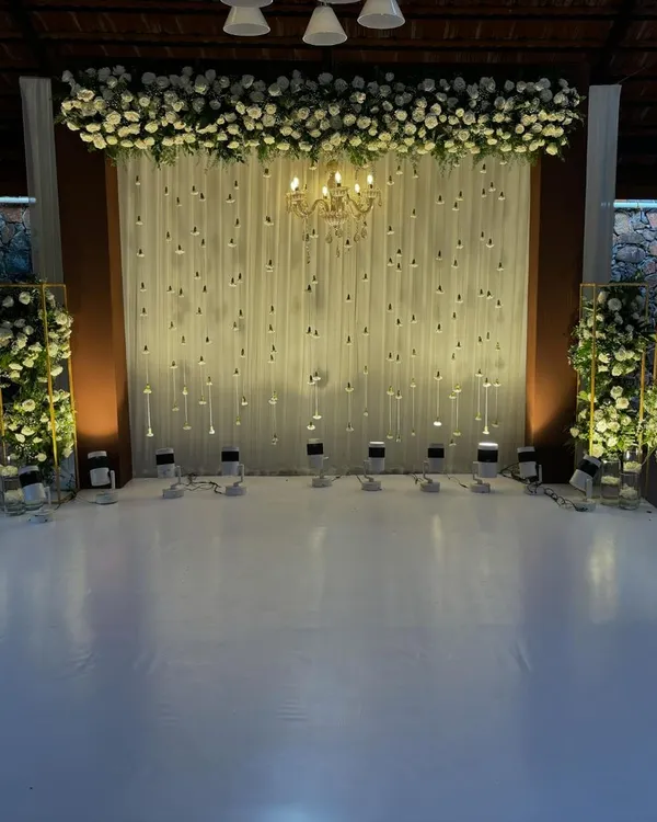 This cozy and elegant setup is perfect for an engagement or reception. The backdrop features a white drape with hanging jasmine buds, topped with a dense arrangement of white roses and a chandelier.