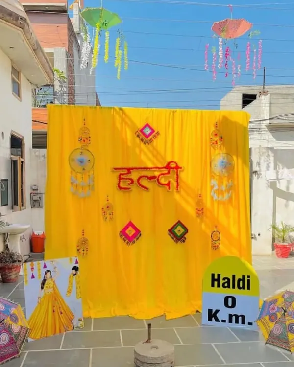 A vibrant outdoor Haldi ceremony backdrop set up on a home terrace. The decor features a bright yellow fabric backdrop with "Haldi" in Hindi, traditional Rajasthani umbrellas, and quirky props like a "Haldi 0 Km" milestone sign, creating a festive and picture perfect setting.