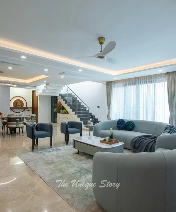 An open-plan living and dining area designed for a modern contemporary home in Jakkur. The space features soft, curved sofas, a sleek marble coffee table, and an elegant 8-seater dining set, all unified by polished marble flooring and sophisticated cove lighting.