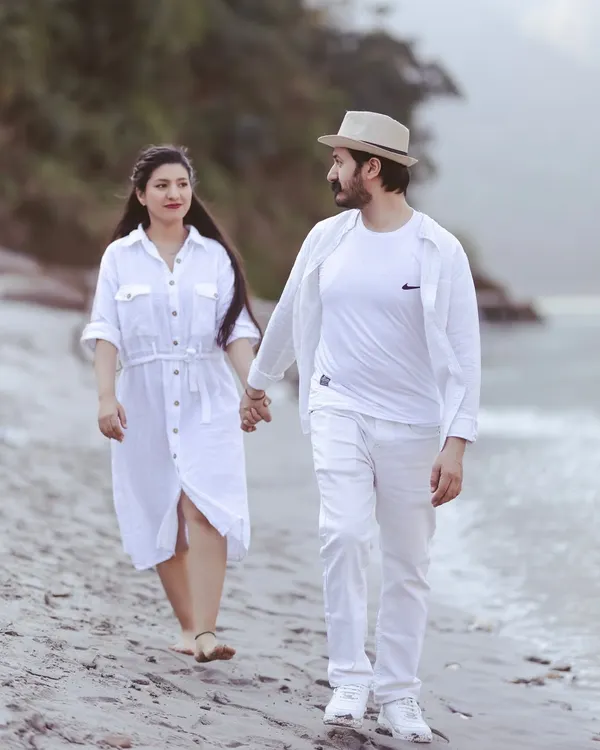 A candid walking shot on the sandy banks of the Ganga. The couple is dressed in white, creating a clean and serene look that complements the natural surroundings.