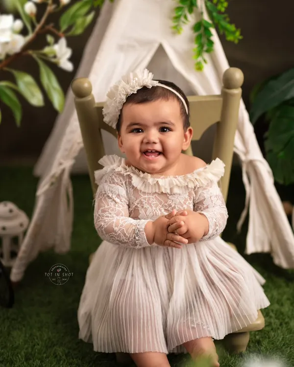A beautiful sitter portrait of a baby girl with a heartwarming smile. Dressed in an elegant white lace outfit, she sits confidently in front of a charming tent backdrop, perfectly capturing this milestone.