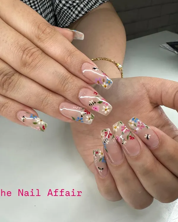 An intricate, hand-painted floral design on long, clear square extensions. This 'meadow' look features tiny flowers and bees for a whimsical, detailed finish.
