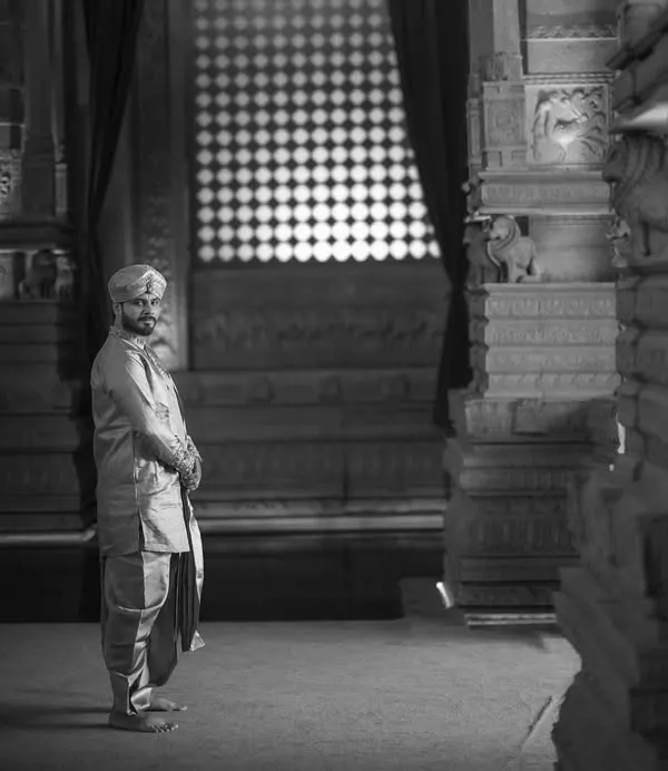 A groom's portrait in black and white, set against the timeless stone pillars of a temple. The dramatic lighting and his solitary figure convey a sense of quiet strength and contemplation.