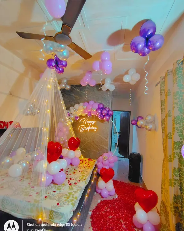 A romantic birthday room setup with a fairy light canopy, a rose petal path, and a purple and white balloon backdrop with a neon sign.