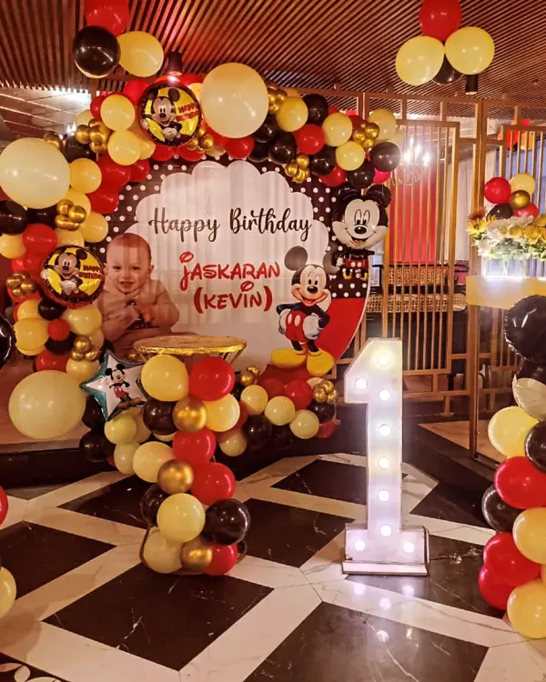 A classic Mickey Mouse theme for a memorable first birthday. This setup at BBQ Company, Rajouri Garden, includes a full balloon arch, character cutouts, and a large light-up number '1' for the perfect photo backdrop.
