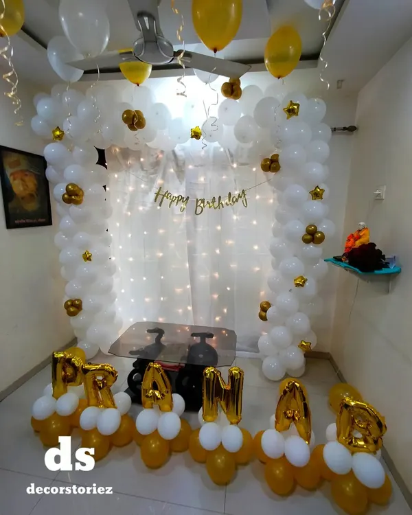An elegant white and gold balloon arch against a fairy light curtain, personalized with the name 'PRANAB' in foil balloons at the base. This is a classic choice for a sophisticated home party.