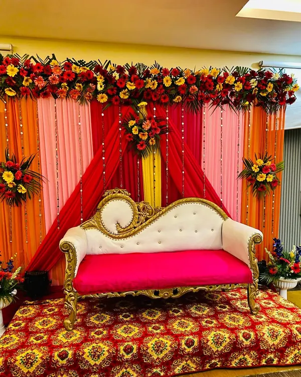 This is a classic wedding stage I set up for a reception in Gurgaon. It features a backdrop of bright red, orange, and pink drapes, a thick garland of fresh gerberas and other flowers, and a traditional white and gold sofa for the couple.